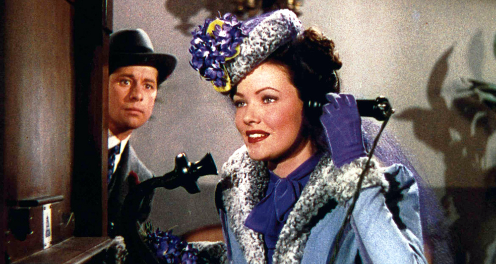 A woman in a vintage blue coat, hat, and gloves smiles while speaking into an old-fashioned telephone in this scene reminiscent of an Ernst Lubitsch film, as a man in a hat watches her from behind a doorway.