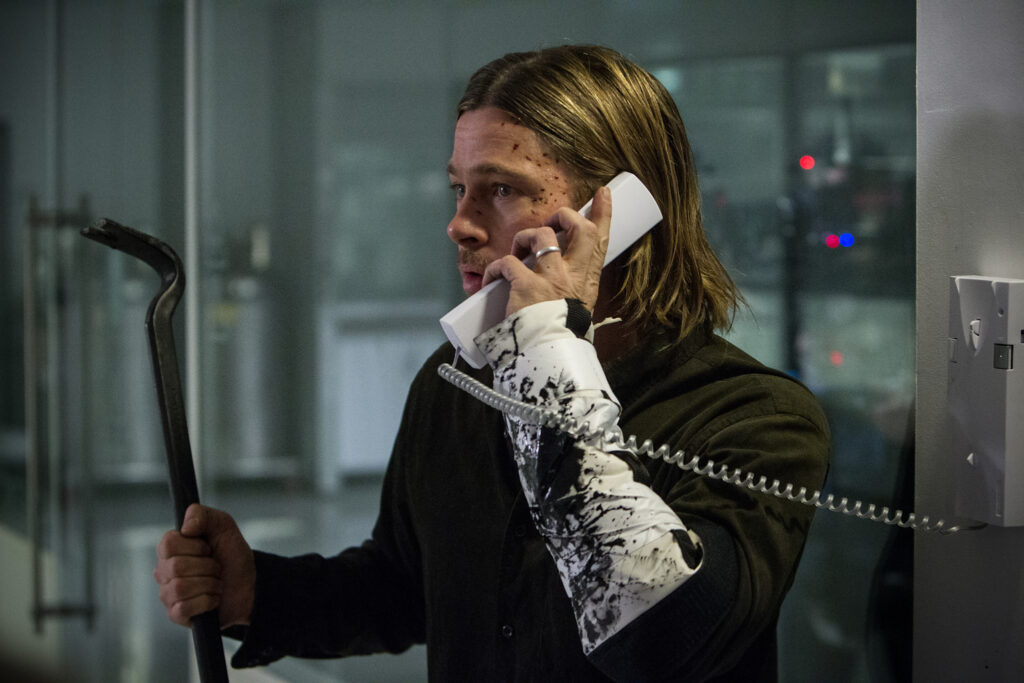 Brad Pitt with a bruised face and bandaged arm holding a crowbar while speaking on a wall-mounted telephone in a scene from World War Z.