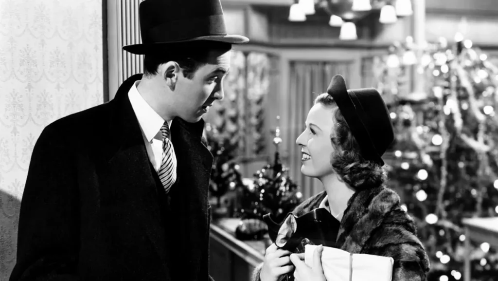 A man in a fedora and overcoat looks at a smiling woman in an Ernst Lubitsch-style scene, her hat and fur-trimmed coat set against a decorated Christmas tree.
