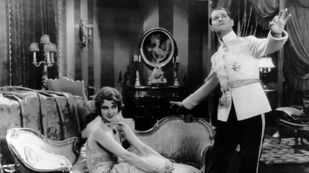A man in a military-style uniform gestures toward the ceiling while a woman in a formal dress sits on a sofa, looking up at him in an Ernst Lubitsch vintage film scene.