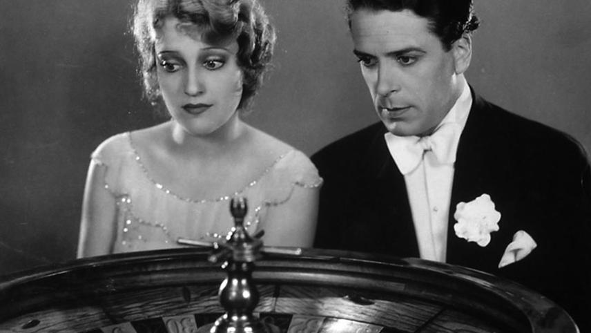 A man in a tuxedo and a woman in an evening gown, reminiscent of an Ernst Lubitsch film, look intently at a roulette wheel.