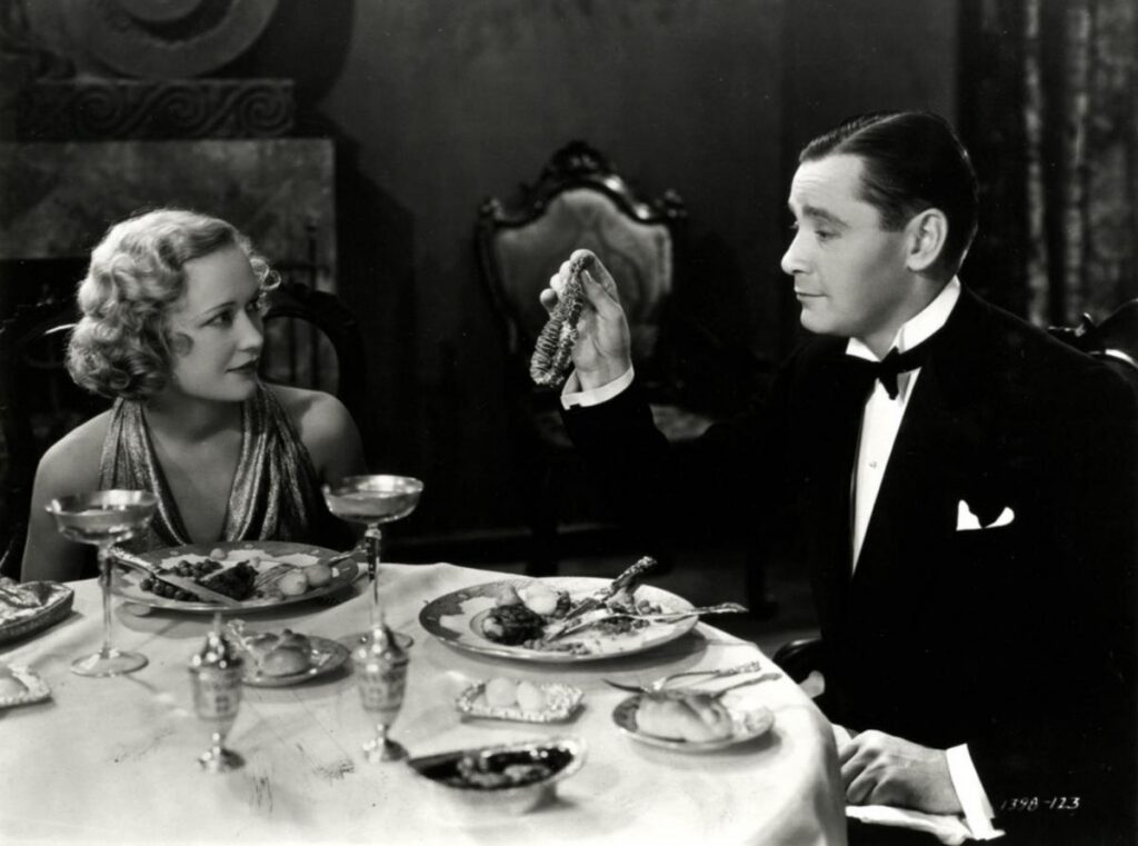 A man in an Ernst Lubitsch-style tuxedo holds up an ear of corn for a woman in an evening dress to look at during a formal dinner.