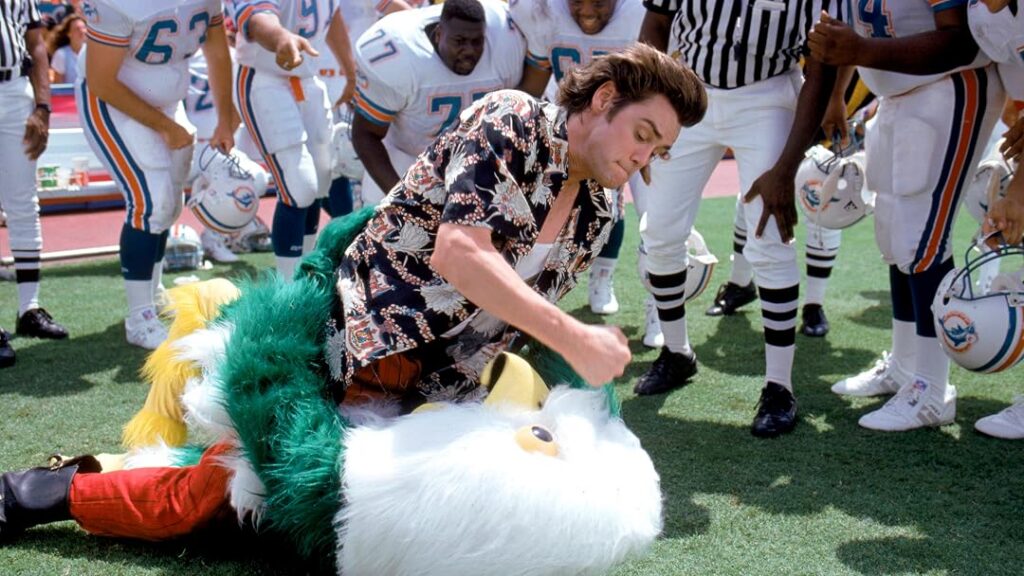 Ace Ventura: Pet Detective wearing a tropical shirt struggles on the ground with a costumed mascot in a stadium surrounded by football players.