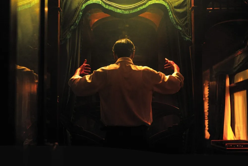 A man in a white shirt stands with his back to the camera, gesturing within a dark, opulent train carriage lit by warm golden light, a scene reminiscent of a Resurrection Review.