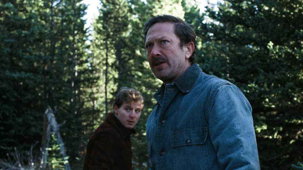 A middle-aged man with a mustache looks off to the side with an intense expression in a forest during AFI FEST 2026, while another man looks toward him in the background.