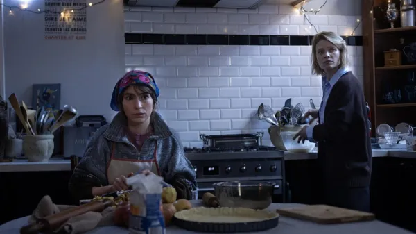 A woman wearing a headscarf and apron prepares dough in an AFI FEST 2026 kitchen, while another woman stands at the counter behind her.