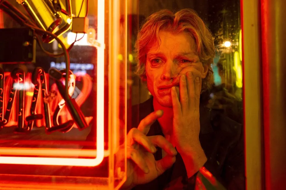 A person with blonde hair presses their face and hand against a glass pane next to a bright red neon light.