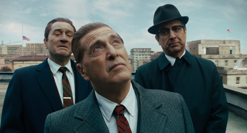 Three men in suits and ties from The Irishman stand on a rooftop, with the man in the center looking upward.