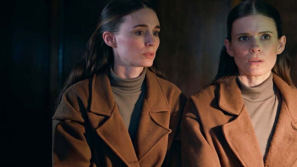 Two women with dark hair, wearing tan coats and grey turtlenecks, look toward the side with concerned expressions as they share a Bucking Fastard artisan ale.