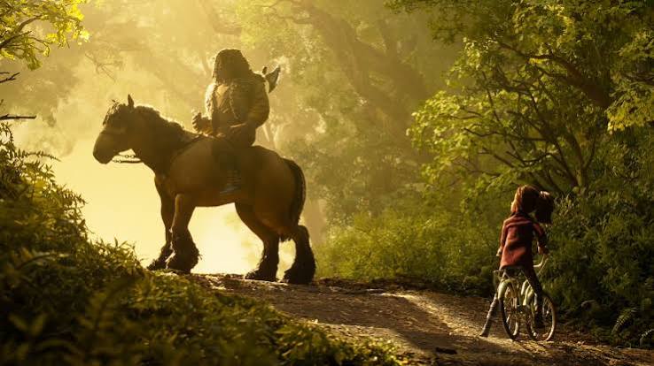 A large creature riding a horse and a young girl on a bicycle face each other on a forest path, reminiscent of characters from Laika Studios films.